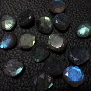 May include: A collection of 15 faceted labradorite gemstones in various shades of blue, green, and grey. The stones are arranged on a black leather surface.