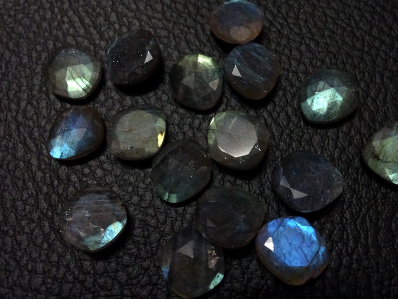 May include: A collection of 15 faceted labradorite gemstones in various shades of blue, green, and grey. The stones are arranged on a black leather surface.