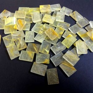 Pode incluir: Uma coleção de pedras de quartzo rutilado dourado retangulares translúcidas. As pedras têm uma cor amarela clara com inclusões douradas.