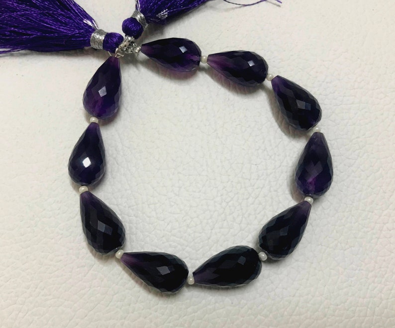May include: A strand of faceted amethyst teardrop beads, ranging in color from deep purple to lighter shades. The beads are strung together with small white spacers. A purple tassel is visible at the top.