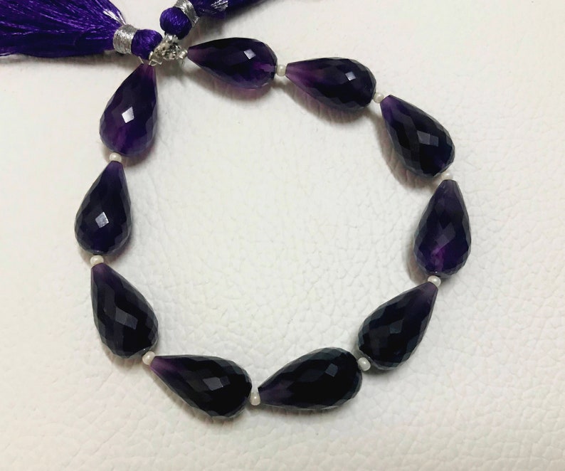 May include: A strand of faceted, teardrop-shaped amethyst beads. The deep purple beads are strung together with small white beads. A purple tassel is visible at the top of the image.