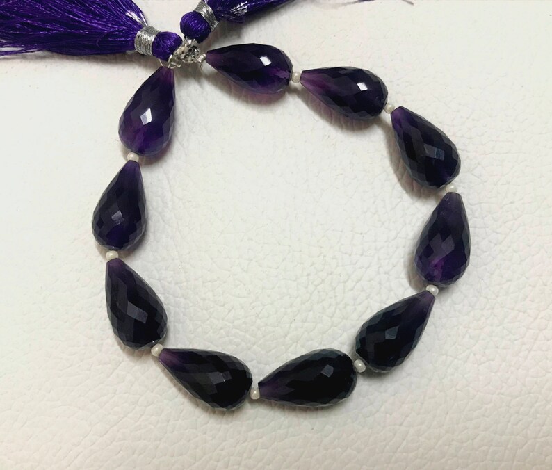 May include: A strand of faceted, teardrop-shaped amethyst beads. The beads are a deep purple color and are strung together with small white beads. A purple tassel is attached to the end of the strand.