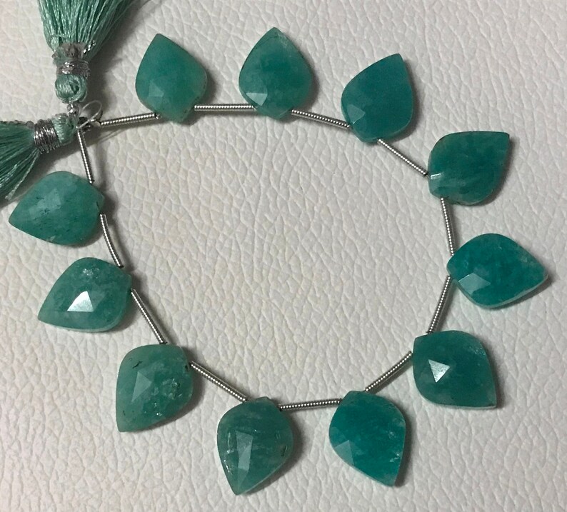 May include: Strand of faceted, teal-colored gemstone beads in a leaf shape. The beads are strung on a thin, silver-colored wire. A matching teal tassel is visible in the upper left corner. The beads are displayed on a white surface.
