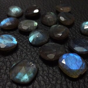 May include: A collection of 14 faceted labradorite gemstones, each with a unique iridescent blue and grey colour. The stones are displayed on a black surface.