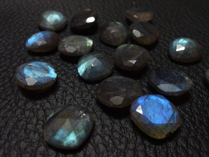 May include: A collection of 14 faceted labradorite gemstones, each with a unique iridescent blue and grey colour. The stones are displayed on a black surface.