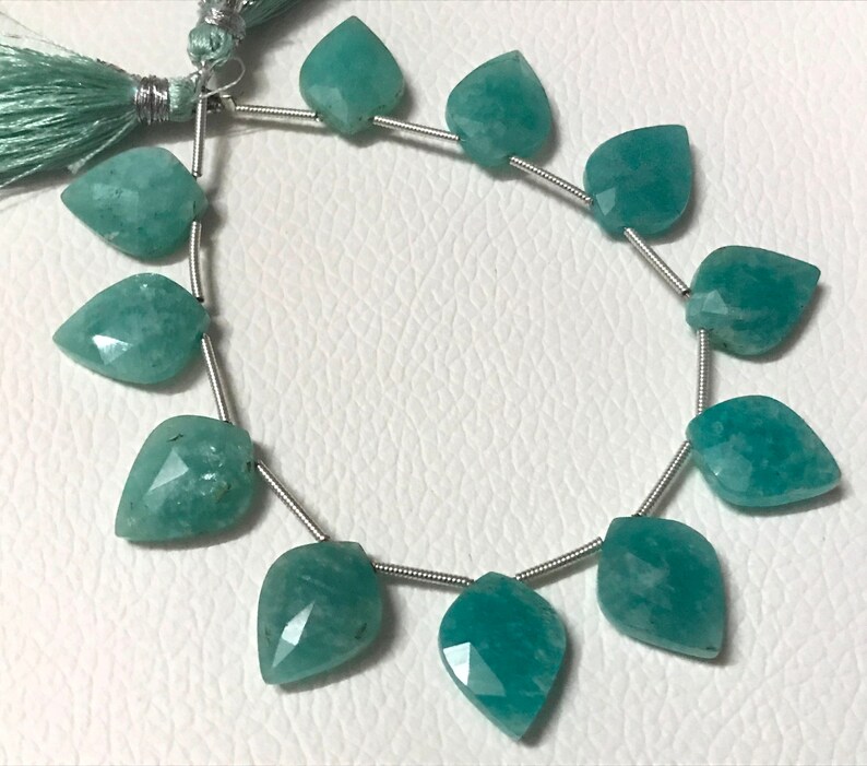 May include: Strand of teal-colored gemstone beads, each shaped like a leaf. The beads are strung on a thin, silver-colored wire. A tassel of teal thread is visible at the top. The beads have a faceted surface.