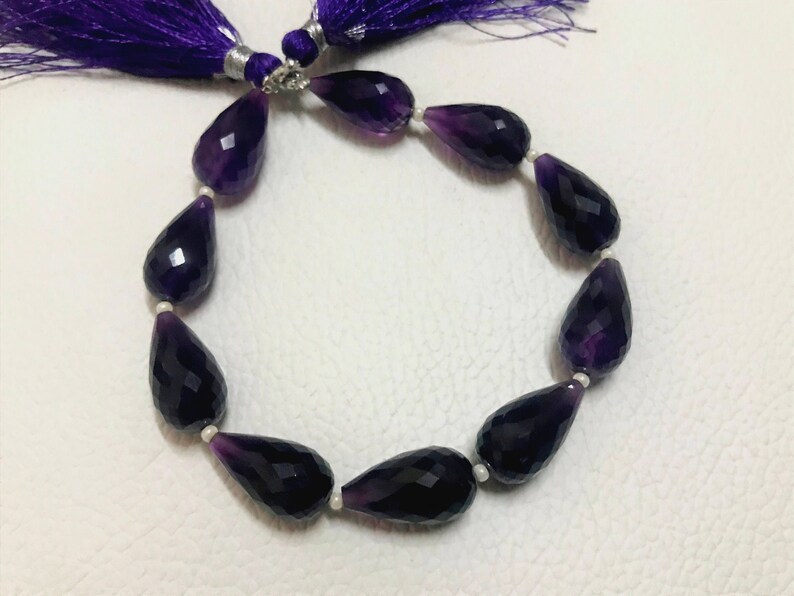 May include: A strand of faceted, teardrop-shaped amethyst beads. The deep purple beads are strung together with small white beads. A purple tassel is attached to the top of the strand.