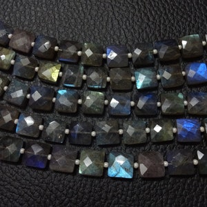 Pode incluir: Um fio de contas de pedra preciosa de labradorita facetada em tons de azul, verde e cinza. As contas são quadradas e têm um acabamento polido.