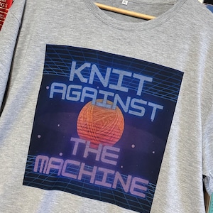 May include: A heather gray XL t-shirt with a graphic that reads "KNIT AGAINST THE MACHINE." The graphic features a ball of yarn in front of a blue and black grid background. The shirt is hanging on a wooden hanger.
