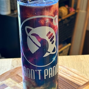 May include: A stainless steel tumbler with a galaxy print and the text "Don't Panic" in a silver colour. The tumbler has a straw and a lid.