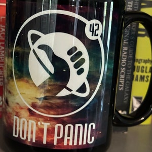 May include: A black ceramic mug with a galaxy print and the text "DON'T PANIC" in white. The mug also features a white spaceship logo with the number 42.