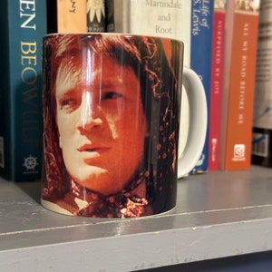 May include: A white ceramic mug featuring a portrait of a person with a light complexion. The mug is surrounded by books on a shelf. The books have titles such as "Tolkien Beowulf" and "A Life of C.S. Lewis".