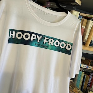 Può includere: T-shirt bianca con la scritta "HOOPY FROOD" in lettere bianche e in grassetto su sfondo blu scuro e turchese. Sotto, la scritta "DON'T PANIC" è in lettere bianche più piccole. La maglietta è appesa a una gruccia nera.