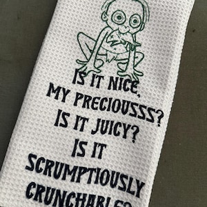 Puede incluir: Pa&ntilde;o de cocina blanco con tejido de gofre con un personaje de dibujos animados verde y el texto "IS IT NICE, MY PRECIOUSSS? IS IT JUICY? IS IT SCRUMPTIOUSLY CRUNCHABLE?" en azul oscuro. La toalla est&aacute; doblada.