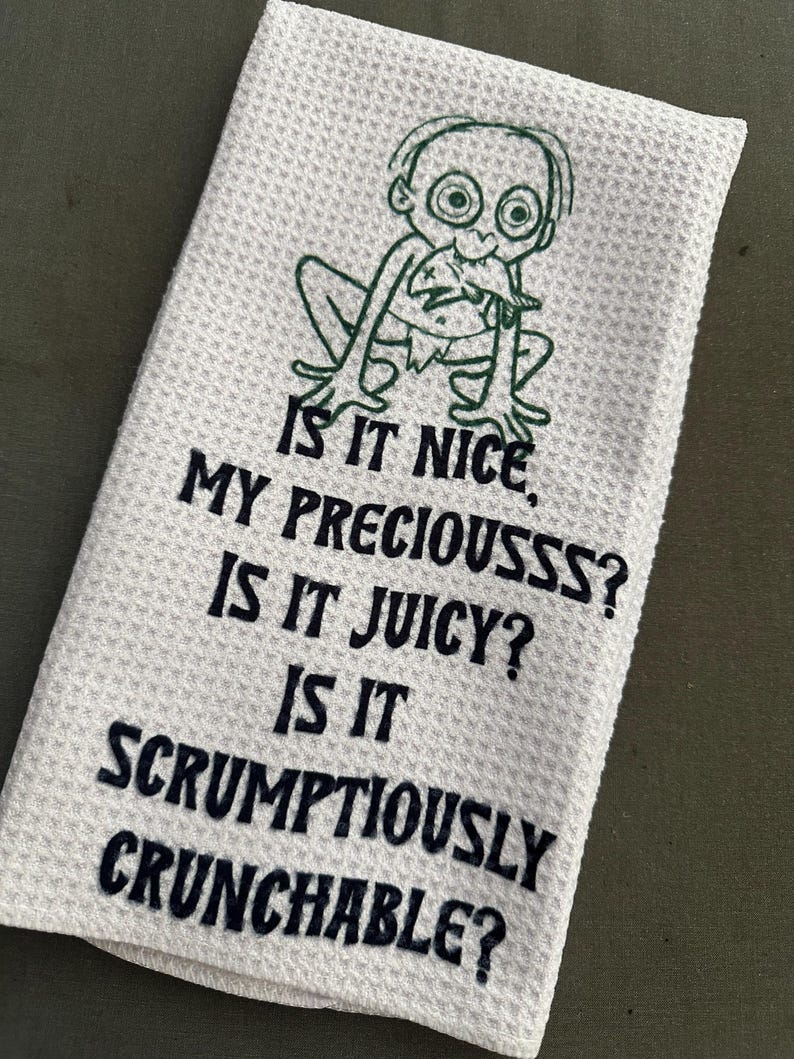 Puede incluir: Pa&ntilde;o de cocina blanco con tejido de gofre con un personaje de dibujos animados verde y el texto "IS IT NICE, MY PRECIOUSSS? IS IT JUICY? IS IT SCRUMPTIOUSLY CRUNCHABLE?" en azul oscuro. La toalla est&aacute; doblada.