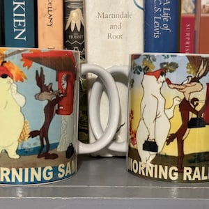 May include: Two white ceramic mugs with cartoon images and text. The mug on the left has a cartoon image of a coyote and a bear with the text "MORNING SAM". The mug on the right has a cartoon image of a coyote and a bear with the text "MORNING RALPH".