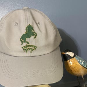 Könnte beinhalten: Eine graue Baseballkappe mit einem grünen galoppierenden Pony-Logo und dem Text "Prancing Pony".