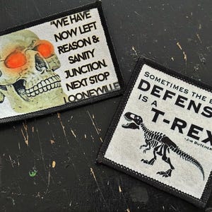 May include: Two embroidered patches. One features a skull with orange eye sockets and the text "WE HAVE NOW LEFT REASON & SANITY JUNCTION NEXT STOP LOONEYVILLE." The other shows a T-Rex skeleton and the text "SOMETIMES THE BEST DEFENSE IS A T-REX - JIM BUTCHER."