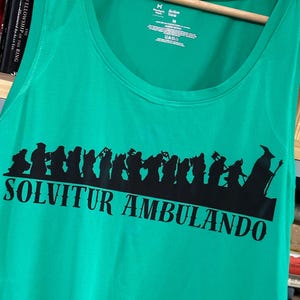 May include: Emerald green vest top featuring a black silhouette of figures and the Latin phrase "SOLVITUR AMBULANDO." The garment is suspended on a wooden hanger. The tag reads "Active Tank".