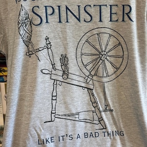 Könnte beinhalten: Graues T-Shirt mit einer schwarz-weißen Illustration eines Spinnrads. Der Text auf dem Shirt lautet: "YOU SAY SPINSTERS LIKE IT'S A BAD THING."