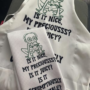 Puede incluir: Delantal blanco y pa&ntilde;o de cocina a juego con un personaje de dibujos animados y las frases "Is it nice, my preciousss? Is it juicy? Is it scrumptiously crunchable?" El dise&ntilde;o es en negro y verde.