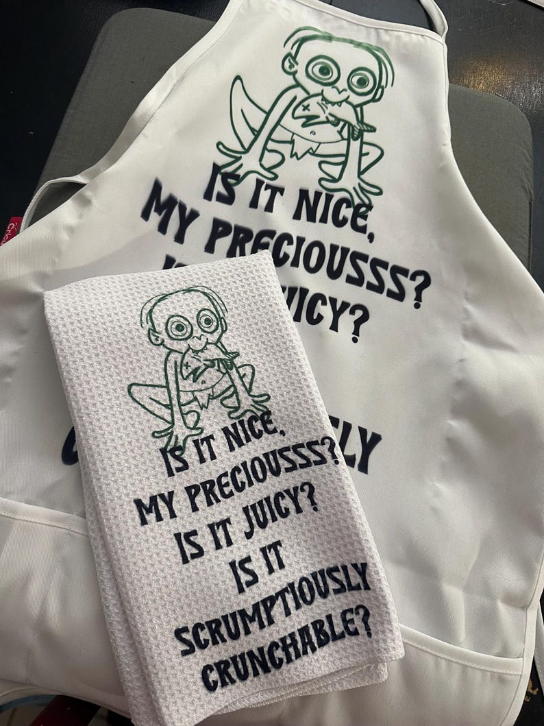Puede incluir: Delantal blanco y pa&ntilde;o de cocina a juego con un personaje de dibujos animados y las frases "Is it nice, my preciousss? Is it juicy? Is it scrumptiously crunchable?" El dise&ntilde;o es en negro y verde.