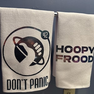 Könnte beinhalten: Zwei cremefarbene Waffelhandtücher hängen an einer Chromstange. Eines zeigt eine schwarze und galaktische Grafik mit dem Text "DON'T PANIC" und der Zahl "42". Das andere zeigt "HOOPY FROOD" in Galaxiefarben.