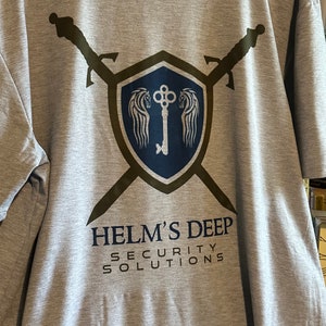 May include: A gray t-shirt featuring a shield design with a key and wings, crossed swords, and the text "HELM'S DEEP SECURITY SOLUTIONS" in navy blue. The design is printed on the front of the shirt.