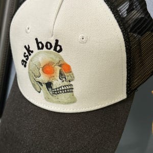 Könnte beinhalten: Schwarz-weiße Trucker-Mütze mit einem Totenkopf-Motiv und dem Text "ask bob". Der Schädel hat orangefarbene Augen. Die Mütze hat eine weiße Vorderseite und eine schwarze Mesh-Rückseite.