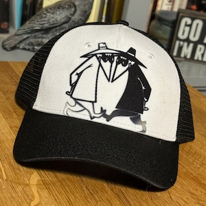 Puede incluir: Una gorra de camionero blanca y negra con una malla negra en la parte posterior. La parte delantera de la gorra presenta una ilustración en blanco y negro de dos figuras que llevan máscaras y sombreros.