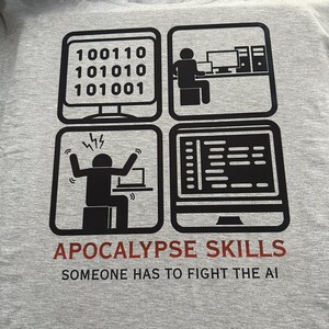 Peut inclure: Un t-shirt gris avec un motif noir et rouge. Le motif présente quatre illustrations sur le thème de l'ordinateur et le texte "APOCALYPSE SKILLS" et "SOMEONE HAS TO FIGHT THE AI". Le t-shirt a un col rond.
