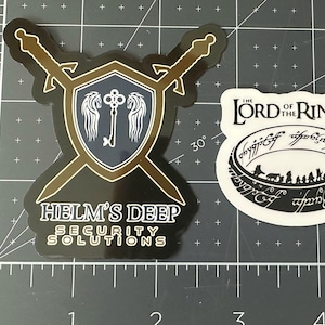 May include: Two stickers, one black with a gold shield and two swords, the text "Helm's Deep Security Solutions", and the other white with the text "The Lord of the Rings" and a black ring with an inscription.