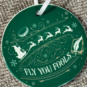 May include: Round, green ceramic ornament with a white ribbon for hanging. The ornament features a design with reindeer pulling a sleigh, snowflakes, and the text "FLY YOU FOOLS" in white and gold.