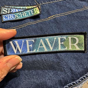 May include: Rectangular fabric patches with black borders and text. One patch reads "WEAVER" in white letters against a blue and green gradient background. Two smaller patches read "SPINNER" and "CROCHETER". The patches are on a denim background.
