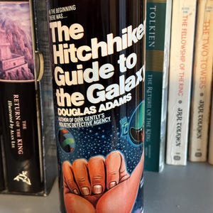 May include: A white tumbler with a black background and a colorful image of a hand giving a thumbs up. The text on the tumbler reads "The Hitchhiker's Guide to the Galaxy" and "Douglas Adams".