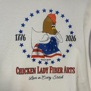 May include: A white t-shirt featuring a graphic of a brown chicken sewing a flag with red and white stripes and blue stars. The design includes the text 1776, 2026, NESTY ROSS, CHICKEN LADY FIBER ARTS, and Love in Every Stitch, all encircled by blue stars.
