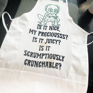 Puede incluir: Delantal blanco con un personaje de dibujos animados verde y el texto "IS IT NICE, MY PRECIOUSSS? IS IT JUICY? IS IT SCRUMPTIOUSLY CRUNCHABLE?" El delantal tiene un bolsillo y lazos en el cuello y la cintura. El delantal es de tela ligera.