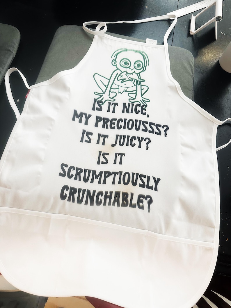 Puede incluir: Delantal blanco con un personaje de dibujos animados verde y el texto "IS IT NICE, MY PRECIOUSSS? IS IT JUICY? IS IT SCRUMPTIOUSLY CRUNCHABLE?" El delantal tiene un bolsillo y lazos en el cuello y la cintura. El delantal es de tela ligera.