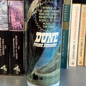 May include: A white travel mug with a black and white image of a desert scene and the text "DUNE FRANK HERBERT" on the front. The text "WINNER OF THE HUGO AWARD AND THE NEBULA AWARD FOR BEST SCIENCE FICTION NOVEL OF THE YEAR!" is also on the front of the mug.