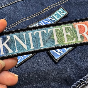 May include: Embroidered patches with the word "KNITTER" in white block letters, set against a colorful gradient background of blue, green, and orange. The patches have a black border and are on a denim background.