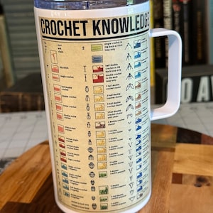May include: White travel mug with a black and red graphic of crochet stitches and their names. The mug has a black handle and a blue lid. The text "CROCHET KNOWLEDGE" is printed in black at the top of the mug.