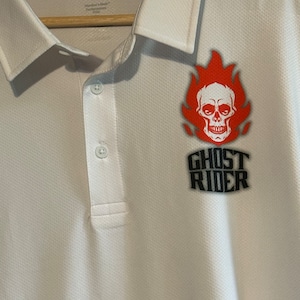 May include: White polo shirt with a black and red Ghost Rider logo featuring a skull with flames. The text "Ghost Rider" is printed below the skull.