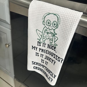 Puede incluir: Toalla de cocina blanca con un personaje de dibujos animados verde y el texto "IS IT NICE, MY PRECIOUSSS? IS IT JUICY? IS IT SCRUMPTIOUSLY CRUNCHABLE?" La toalla est&aacute; colgada de un tirador de horno.