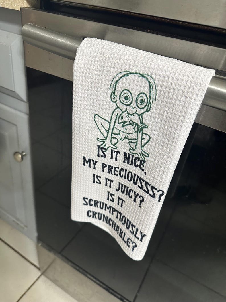 Puede incluir: Toalla de cocina blanca con un personaje de dibujos animados verde y el texto "IS IT NICE, MY PRECIOUSSS? IS IT JUICY? IS IT SCRUMPTIOUSLY CRUNCHABLE?" La toalla est&aacute; colgada de un tirador de horno.