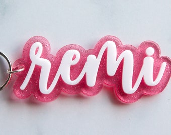 May include: A pink glitter keychain with the name "remi" in white cursive lettering. The keychain has a silver metal ring attached. The name is in a cloud-like shape.