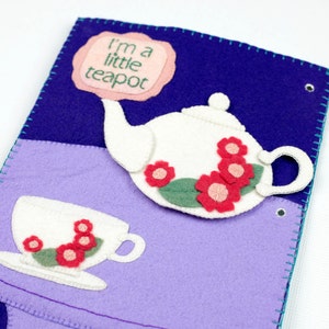 Può includere: Un libro di feltro viola e blu con una teiera e una tazza bianche. La teiera ha fiori rosa e foglie verdi. Il libro ha il testo "I'm a little teapot" sulla copertina.