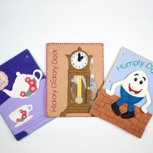 May include: Three felt board books for children. The first book is purple and features a teapot with the text "I'm a little teapot". The second book is brown and features a clock with the text "Hickory Dickory Dock". The third book is blue and features a smiling egg with the text "Humpty Dumpty".