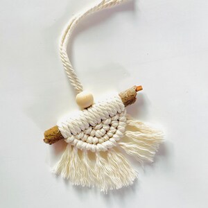 May include: A white macrame wall hanging with a wooden branch and a wooden bead. The macrame has a fringe at the bottom.