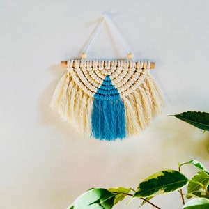 May include: A small, handmade macrame wall hanging with a cream and blue color scheme. The hanging features a geometric pattern with fringe details.
