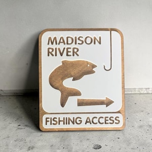 May include: A rectangular wooden sign with a white face featuring the text MADISON RIVER and FISHING ACCESS in brown. The sign includes a brown cutout graphic of a fish, a fishing hook, and a directional arrow pointing to the right side.
